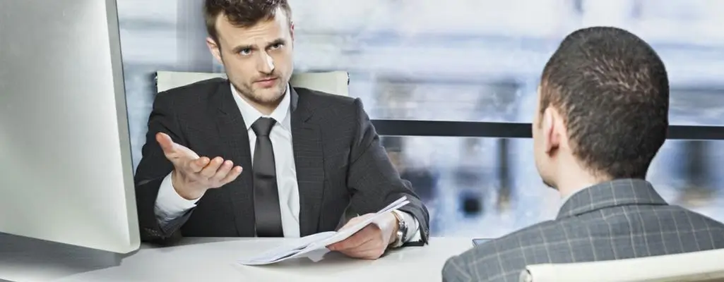 Zdjęcie w artykule - co daje orzeczenie o niepełnosprawności pracodawcy. Scena w biurze. Mężczyzna w garniturze trzymający w ręce dokument tłumaczy coś siedzącemu na przeciw niego drugiemu mężczyźnie. Obok na biurku stoi komputer.