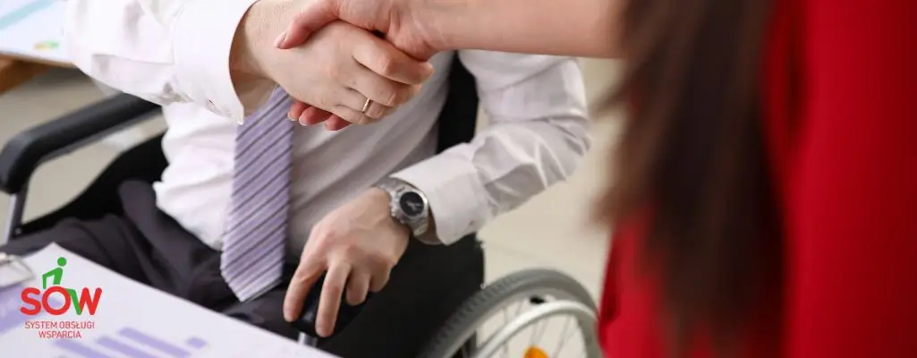 Obrazek wyróżniający w artykule - sow pfron. Na zdjęciu widzimy mężczyznę na wózku inwalidzkim oraz kobietę stojącą obok niego. Kobieta, podaje rękę w geście potwierdzenia umowy. Na dokumentach obok komputera widnieje logo "Systemu Obsługi Wsparcia" (SOW).