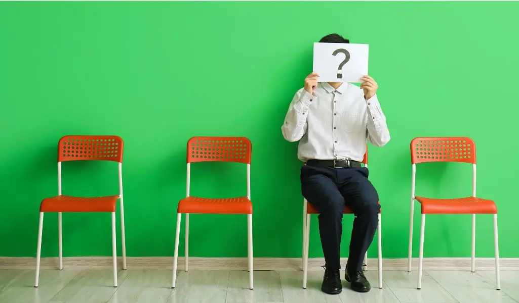 Obrazek wyróżniający w artykule - dlaczego chcesz pracować w naszej firmie. Obraz przedstawia mężczyznę siedzącego na jednym z trzech pomarańczowych krzeseł na tle jasnej, zielonej ściany. Mężczyzna trzyma przed twarzą kartkę z dużym znakiem zapytania. Obraz może symbolizować oczekiwanie, anonimowość, niepewność lub pytania dotyczące przyszłości, na przykład w kontekście poszukiwania pracy lub rekrutacji.