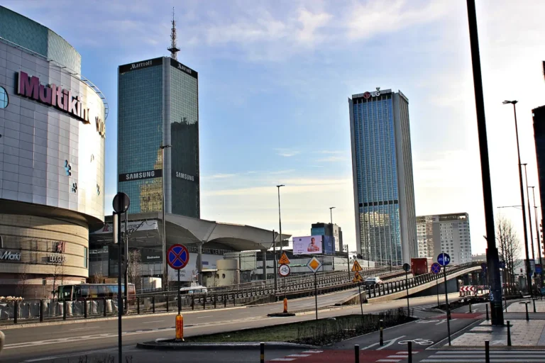 Zdjęcie przedstawia miejski krajobraz Warszawy, ze znanymi wieżowcami w tle. Po lewej stronie widoczny jest budynek Multikina Złote Tarasy, którego fasada pokryta jest nowoczesnym szkłem i metalem, a na niej umieszczone są duże napisy reklamowe, w tym logo H&M oraz innych marek. W centrum zdjęcia, za budynkiem Multikina, znajduje się charakterystyczny wieżowiec z napisem "Marriott" na szczycie oraz reklamami Samsunga na kilku piętrach. Po prawej stronie widoczny jest inny wysoki budynek z logo LG na szczycie. Na pierwszym planie zdjęcia widoczna jest ulica z oznaczeniami drogowymi, w tym znakiem zakazu parkowania oraz znakami ostrzegawczymi dotyczącymi rowerzystów i pieszych. W tle widoczny jest wiadukt drogowy prowadzący do Warszawy Centralnej, głównego dworca kolejowego w mieście. Cała scena rozgrywa się w pogodny dzień, z jasnym, niebieskim niebem na horyzoncie.