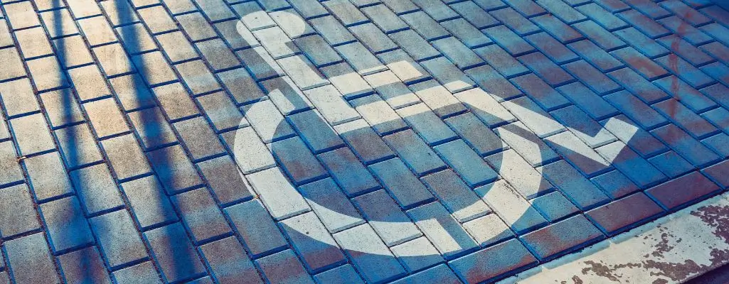 Zdjęcie przedstawia miejsce parkingowe dla niepełnosprawnych, oznaczone charakterystycznym białym symbolem wózka inwalidzkiego. Nawierzchnia parkingu jest wykonana z kostki brukowej w odcieniach niebieskiego i szarego. Symbol wózka inwalidzkiego jest wyraźnie widoczny na tle niebieskiej kostki brukowej. Widać również cienie padające na powierzchnię parkingu, co sugeruje, że zdjęcie zostało zrobione w ciągu dnia przy słonecznej pogodzie.