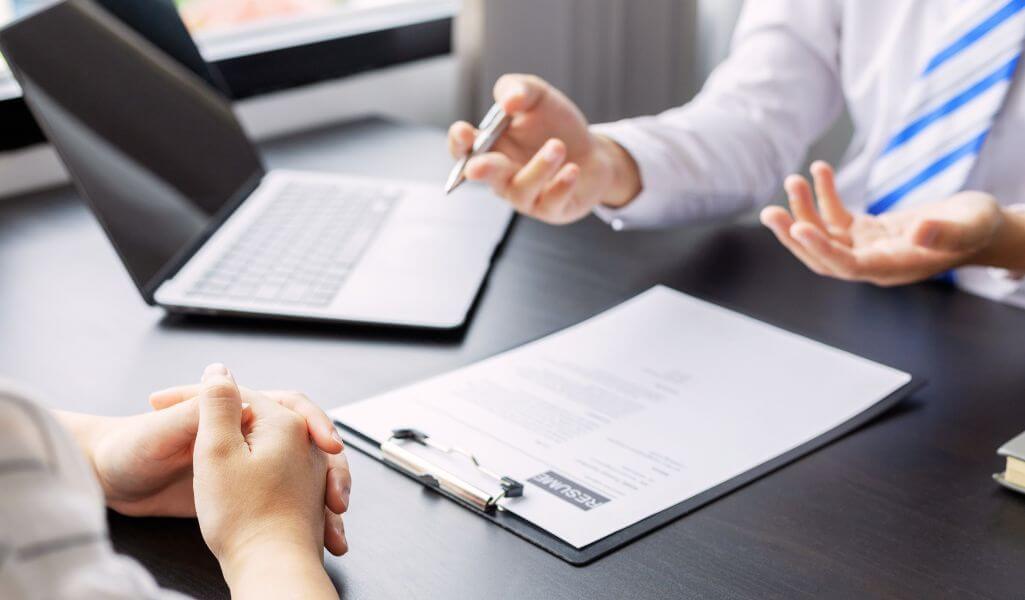 Obrazek wyróżniający w artykule - słabe strony podczas rozmowy kwalifikacyjnej. Na obrazie widoczne są dwie osoby siedzące naprzeciwko siebie przy stole, co sugeruje, że znajdują się one w trakcie rozmowy kwalifikacyjnej lub spotkania biznesowego. Na pierwszym planie widać dłonie jednej osoby, złożone na stole, podczas gdy druga osoba gestykuluje, trzymając długopis. Na stole leży otwarta teczka z dokumentem oznaczonym słowem „RESUME” (ang. CV), co dodatkowo wskazuje na kontekst rozmowy kwalifikacyjnej. W tle znajduje się otwarty laptop. Cała scena jest dobrze oświetlona, co sugeruje, że pokój ma dostęp do naturalnego światła.