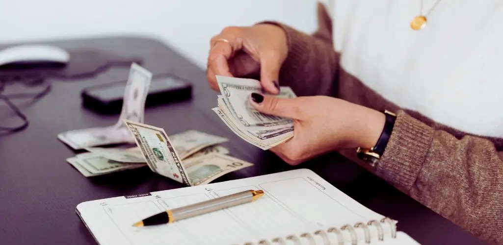 Obrazek wyróżniający w artykule - ile pieniędzy przysługuje osobie z grupą. Obrazek przedstawia osobę siedzącą przy biurku, przeliczającą plik banknotów. Banknoty są w różnych nominałach, a waluta to dolary amerykańskie. Na biurku znajduje się otwarty notes, obok którego leży długopis. W tle, poza ostrością, można zauważyć telefon oraz inne banknoty. Osoba nosi bransoletkę oraz naszyjnik z zawieszką, a jej ubiór to sweter w odcieniach brązu i beżu. Scena sugeruje zajmowanie się finansami, budżetem lub innymi działaniami związanymi z przeliczaniem pieniędzy.