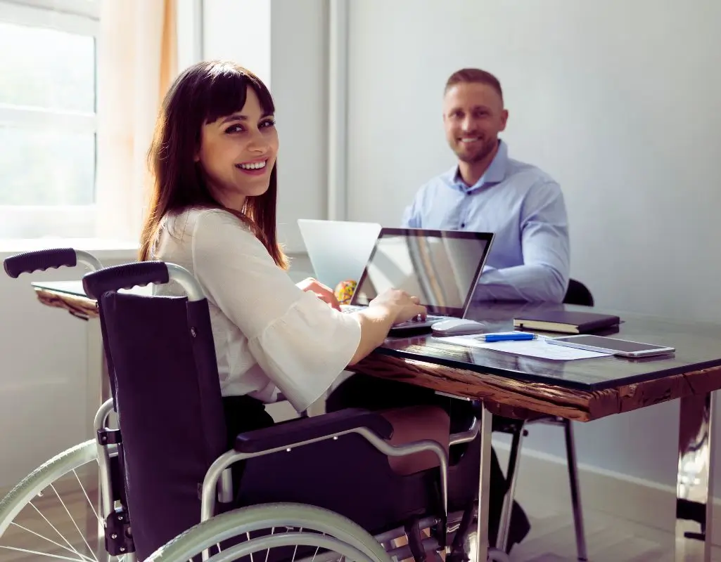 Obrazek wyróżniający w artykule - uprawnienia pracowników z orzeczeniem. Na obrazku widzimy kobietę na wózku inwalidzkim, siedzącą przy biurku z laptopem. Uśmiecha się i patrzy w stronę kamery, co nadaje zdjęciu przyjazny i pozytywny charakter. Przy biurku siedzi także mężczyzna, również uśmiechnięty, co sugeruje przyjazną atmosferę pracy. Na biurku znajdują się różne przedmioty, takie jak długopis, notes i smartfon, co wskazuje na profesjonalne środowisko biurowe. Obraz oddaje inkluzywność oraz pozytywne podejście do zatrudniania osób z niepełnosprawnościami.