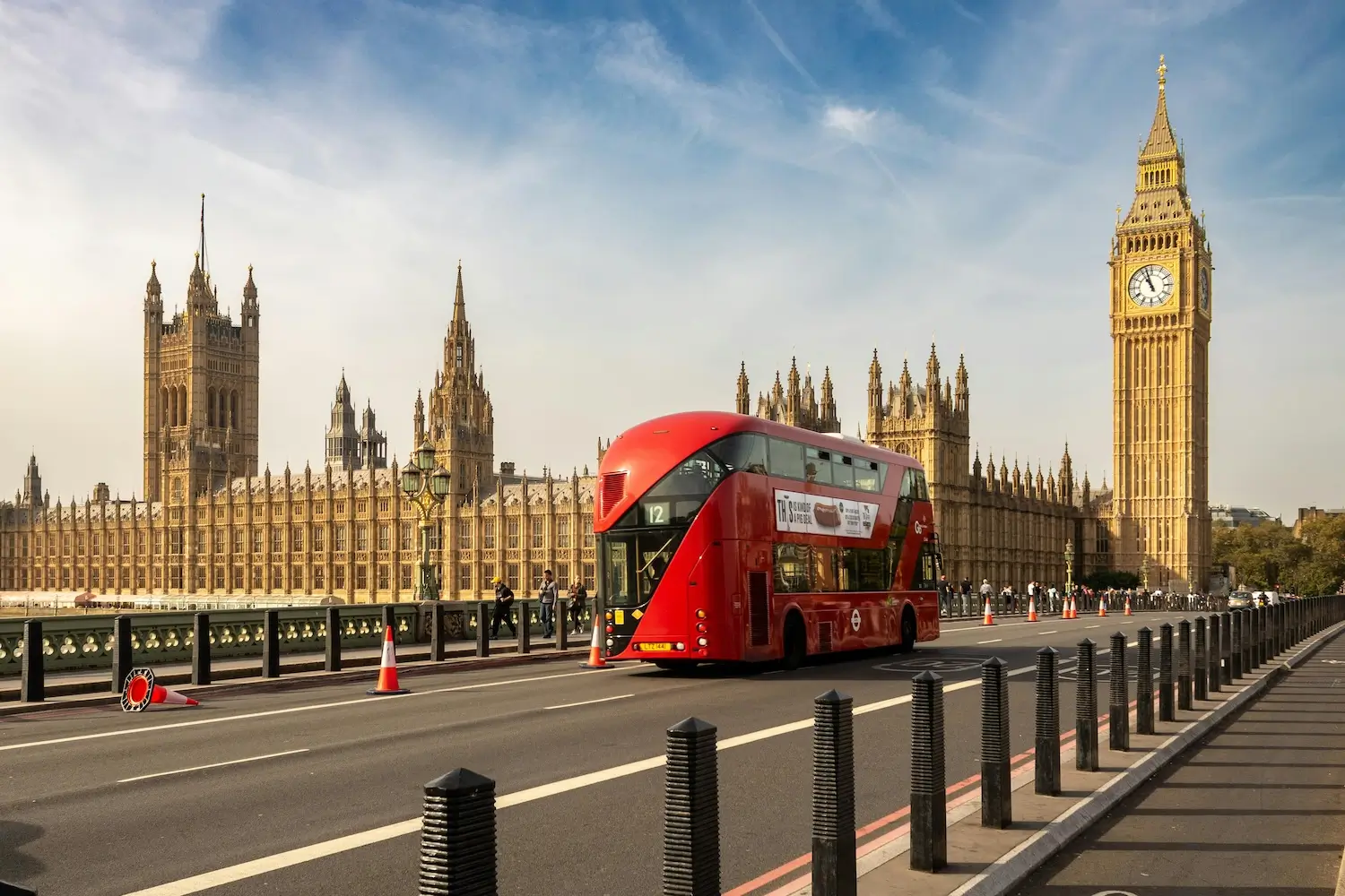 Na zdjęciu widoczny jest jeden z charakterystycznych symboli Londynu — czerwony, piętrowy autobus przejeżdżający przez most Westminster Bridge, na tle budynków parlamentu brytyjskiego oraz wieży zegarowej Big Ben. Jasne, gotyckie budynki parlamentu z licznymi wieżyczkami i detalami architektonicznymi kontrastują z czerwienią autobusu. Niebo jest jasne, z lekkim zachmurzeniem, a na moście widoczne są dodatkowo pachołki ostrzegawcze. Całość oddaje klimat Londynu, znanego z historycznej architektury oraz nowoczesnej, dynamicznej infrastruktury miejskiej.