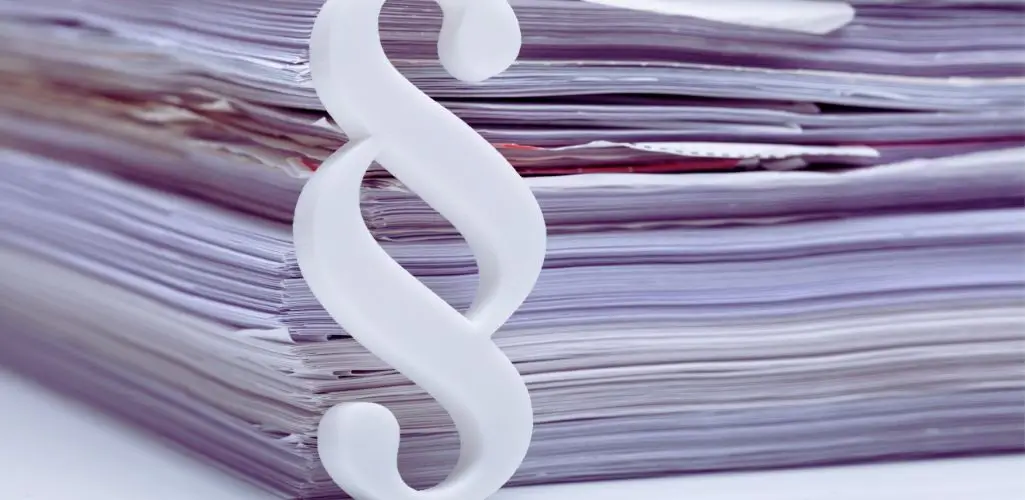 Obrazek wyróżniający w artykule - Orzeczenie o niepełnosprawności. Na zdjęciu znajduje się stos dokumentów, ułożony w równą stertę, obok którego stoi biały symbol paragrafu "§". Dokumenty mają lekko fioletowy odcień, co dodaje zdjęciu chłodnego, formalnego charakteru. Symbol paragrafu jest duży i wyraźny, co sugeruje związek z przepisami prawnymi, regulacjami lub procedurami administracyjnymi dotyczącymi tych dokumentów. Zdjęcie może odnosić się do kwestii prawnych związanych z dokumentacją, takich jak przepisy prawne lub wymagania dokumentacyjne.