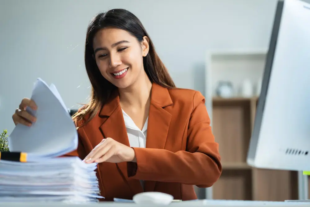Zdjęcie przedstawia kobietę siedzącą przy biurku, zajmującą się przeglądaniem dokumentów. Kobieta ma długie, ciemne włosy i uśmiecha się, co nadaje zdjęciu pozytywnego charakteru. Ubrana jest w elegancki, brązowy żakiet, pod którym widoczna jest biała bluzka. Przed nią leży stos dokumentów, które przegląda, co sugeruje, że jest zaangażowana w pracę biurową, najprawdopodobniej księgowość lub administrację. W tle znajduje się nowoczesne wnętrze biurowe z półkami i komputerem na biurku.
