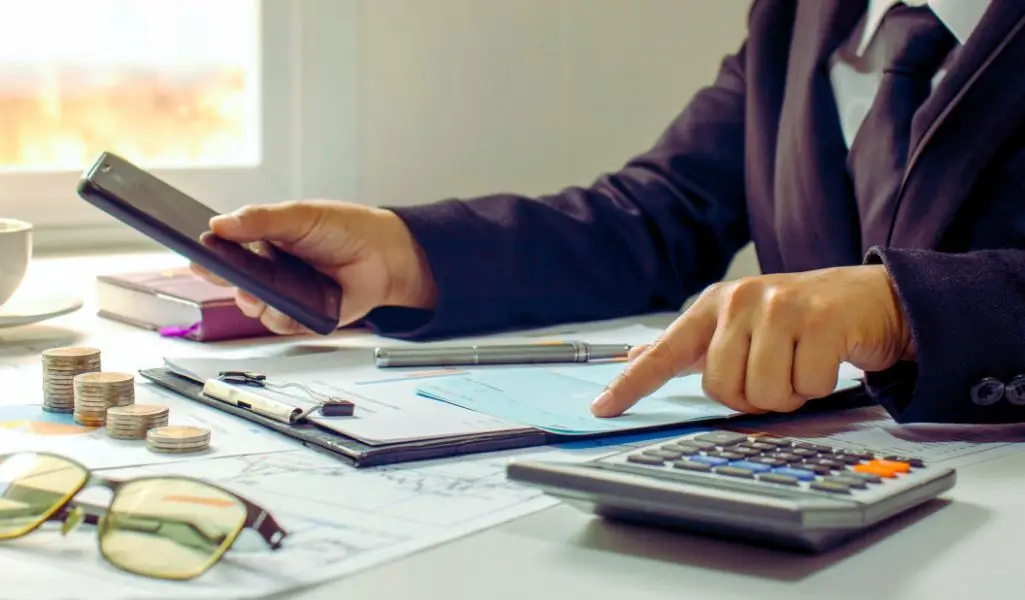 Obrazek do artykułu - warunki otrzymania dofinansowania. Obraz przedstawia osobę w eleganckim stroju biurowym, siedzącą przy biurku. Na biurku widoczne są narzędzia związane z finansami i organizacją: kalkulator, monety ułożone w stosy, dokumenty na klipbordzie, długopis oraz smartfon. Osoba jedną ręką trzyma telefon, a drugą wskazuje coś na dokumencie, co sugeruje dokładne sprawdzanie lub analizowanie danych. Scena oddaje atmosferę skupienia i profesjonalizmu, a także dbałość o szczegóły. Może kojarzyć się z pracą biurową, planowaniem finansowym lub wypełnianiem formalności. W tle widać książki, co podkreśla organizację i wiedzę, a okulary na biurku mogą sugerować potrzebę precyzji i analizy.