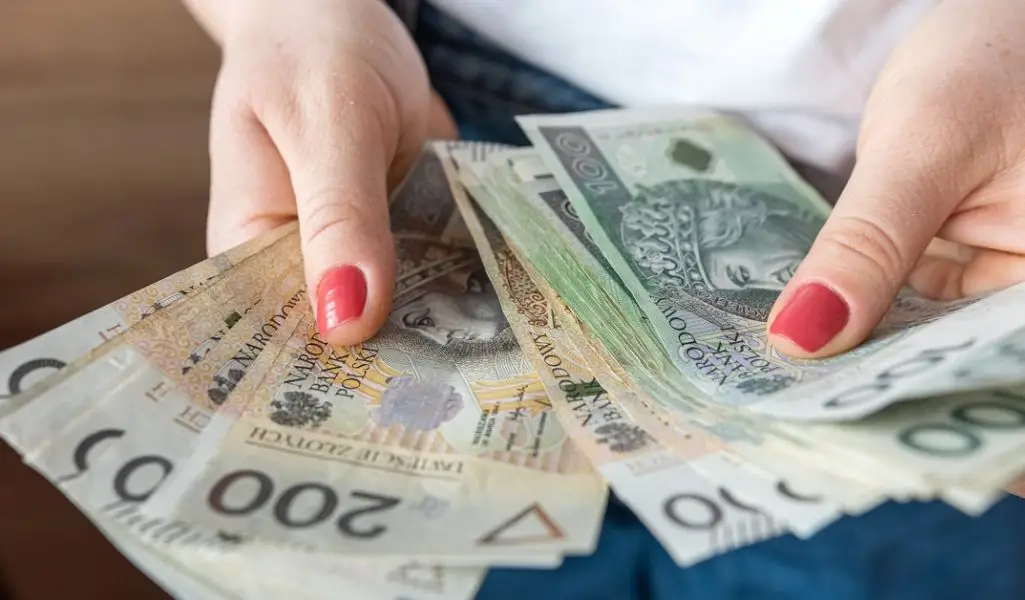 Na zdjęciu widać zbliżenie dłoni kobiety trzymającej wachlarz banknotów polskich złotych – widoczne są nominały 10, 100 i 200 zł. Jej paznokcie są pomalowane na czerwono, a sposób, w jaki trzyma pieniądze, sugeruje pewność, ulgę i może nawet dumę z otrzymanego wsparcia. To obraz, który przywołuje skojarzenia z realną pomocą finansową i poczuciem bezpieczeństwa. Idealnie oddaje moment, w którym „dofinansowanie do ogrzewania z PFRON” staje się czymś namacalnym – pieniędzmi w rękach, które mogą ogrzać dom i życie.