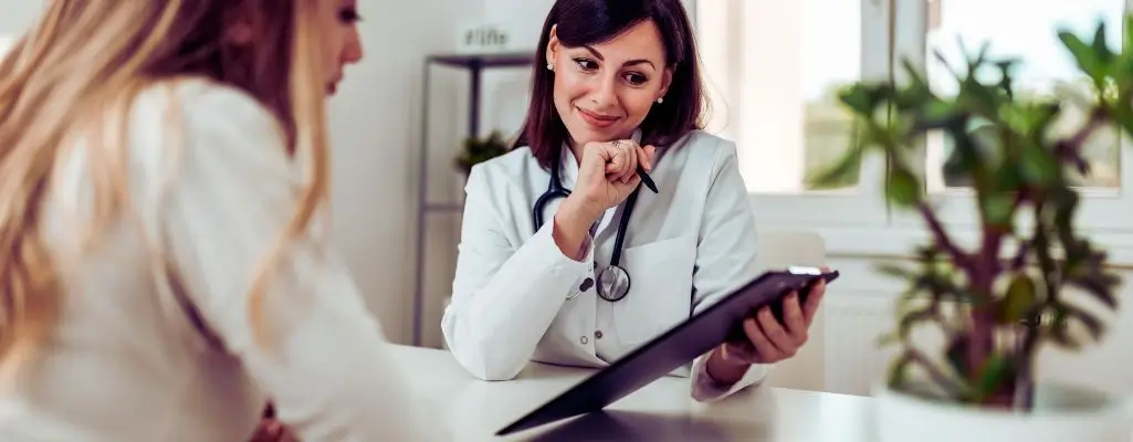 Na obrazku widać lekarkę w białym fartuchu, z uśmiechem i życzliwym spojrzeniem, rozmawiającą z pacjentką. Kobieta trzyma w ręku teczkę z dokumentami i pokazuje coś pacjentce, prawdopodobnie omawiając ważne kwestie zdrowotne. W tle widać jasne pomieszczenie z rośliną doniczkową i regałem. Atmosfera na zdjęciu jest spokojna i pełna zaufania – sugeruje, że pacjentka otrzymuje zrozumiałe i wspierające informacje. Obrazek może ilustrować konsultację dotyczącą możliwości wsparcia, takich jak wykaz sprzętu rehabilitacyjnego refundowanego przez PFRON.