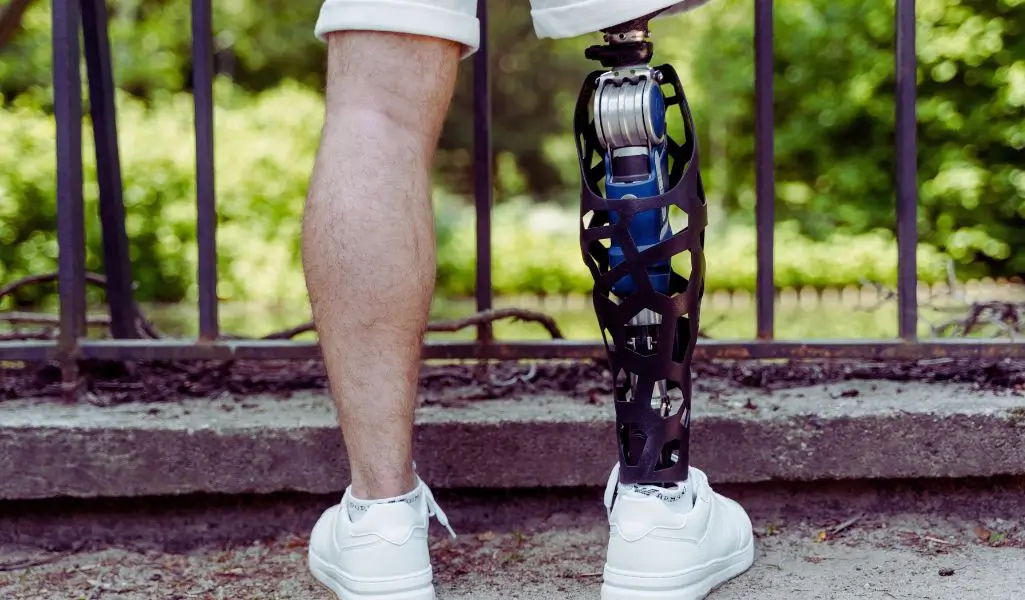Na zdjęciu widzimy mężczyznę stojącego tyłem, ubranego w białe buty sportowe i jasne krótkie spodenki. Jego prawa noga jest naturalna, a lewa zastąpiona nowoczesną protezą o futurystycznym designie – z widocznymi metalowymi elementami i ażurową, czarną osłoną. Tło stanowi park lub ogród z zielenią, a mężczyzna stoi przy metalowym ogrodzeniu, jakby zatrzymał się na chwilę spaceru. Obraz ukazuje nie tylko technologiczną precyzję protezy, ale przede wszystkim siłę i niezależność osoby z niepełnosprawnością. Zdjęcie symbolizuje wolność, aktywność i nową jakość życia, jaką może przynieść dofinansowanie do protezy kończyny z PFRON.
