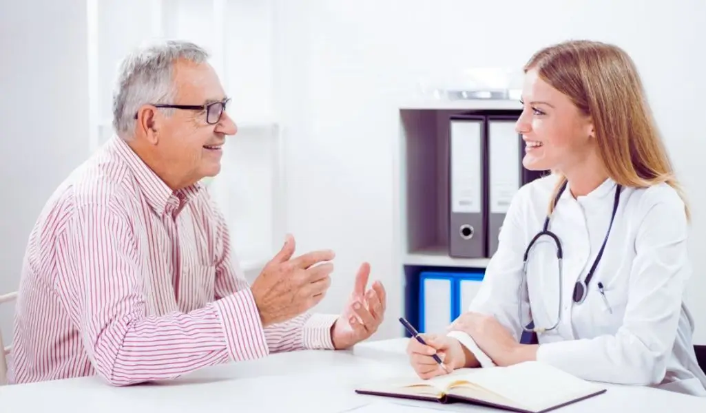 Uśmiechnięty starszy mężczyzna w okularach i koszuli w czerwono-białe paski siedzi przy biurku naprzeciw młodej lekarki w białym kitlu ze stetoskopem; oboje gestykulują i patrzą na siebie z życzliwością, tworząc atmosferę zaufania, ulgi i zrozumienia podczas rozmowy o tym, jak dostać rentę na kręgosłup.