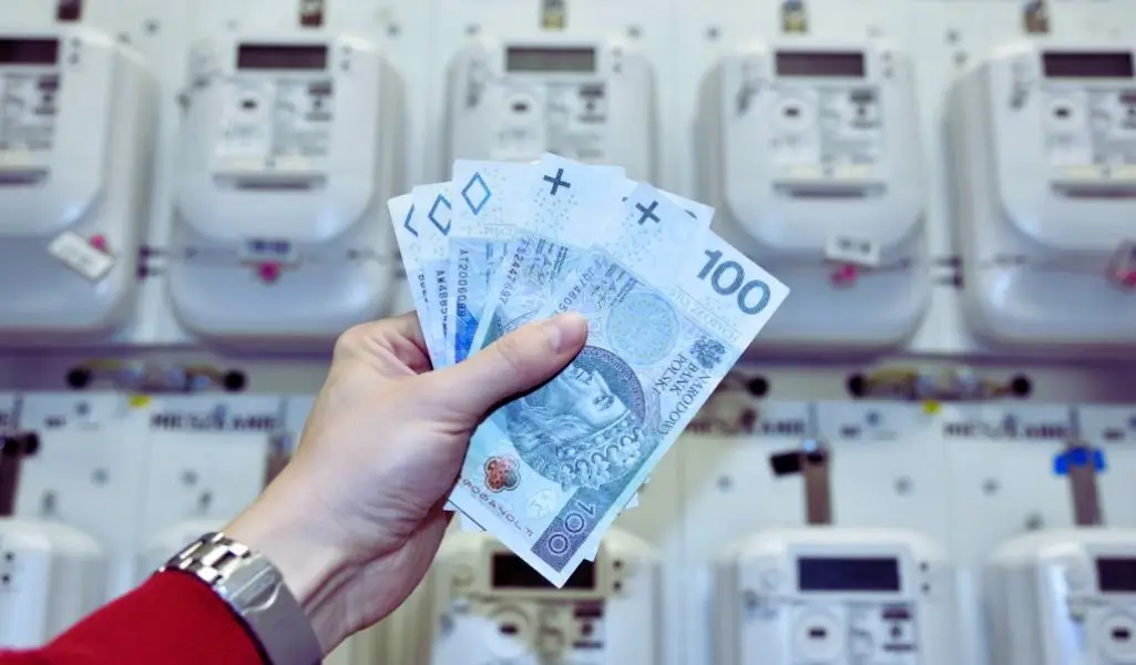 Zdjęcie przedstawia zbliżenie dłoni trzymającej wachlarz banknotów 100 złotowych na tle nowoczesnych liczników energii elektrycznej. Ręka, ubrana w czerwony rękaw i srebrny zegarek, symbolicznie pokazuje pieniądze przeznaczone na opłaty za prąd. Obraz wywołuje skojarzenia z wysokimi kosztami energii, ale też daje nadzieję i ulgę – sugeruje, że możliwe jest wsparcie finansowe. Idealnie oddaje temat „dodatek do energii elektrycznej z PFRON” jako realną pomoc dla osób z niepełnosprawnością w codziennych wydatkach.