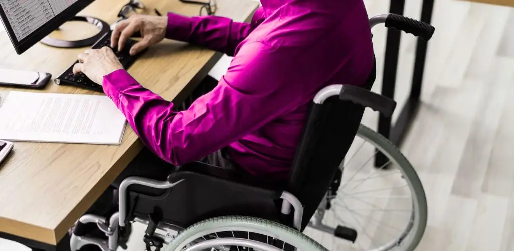 Na zdjęciu widać osobę na wózku inwalidzkim ubraną w elegancką, fuksjową koszulę, pracującą przy biurku z komputerem. Jej ręce spoczywają na klawiaturze, obok leży dokument i telefon – symbole zawodowego zaangażowania i samodzielności. Obraz oddaje atmosferę skupienia, profesjonalizmu i niezależności. Przedstawia codzienność, w której praca biurowa staje się dostępna dzięki elastycznym rozwiązaniom. To wizualna reprezentacja hasła: korzyści pracy zdalnej dla osoby niepełnosprawnej i pracodawcy – pokazująca, że dzięki nowoczesnym technologiom możliwe jest pełne uczestnictwo w życiu zawodowym bez barier.