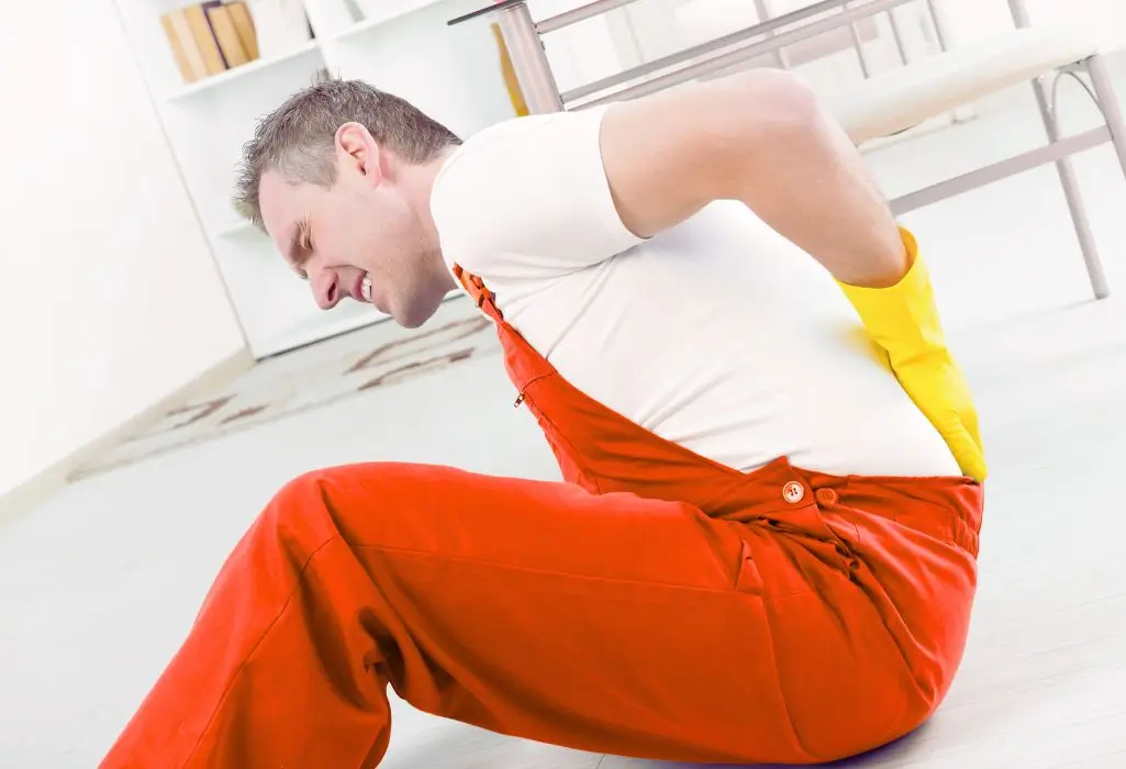 Mężczyzna w roboczym kombinezonie siedzi na podłodze, trzymając się za plecy z wyraźnym grymasem bólu – scena ukazuje moment urazu w pracy, który może mieć wpływ na wysokość odszkodowanie z ZUS za wypadek przy pracy.