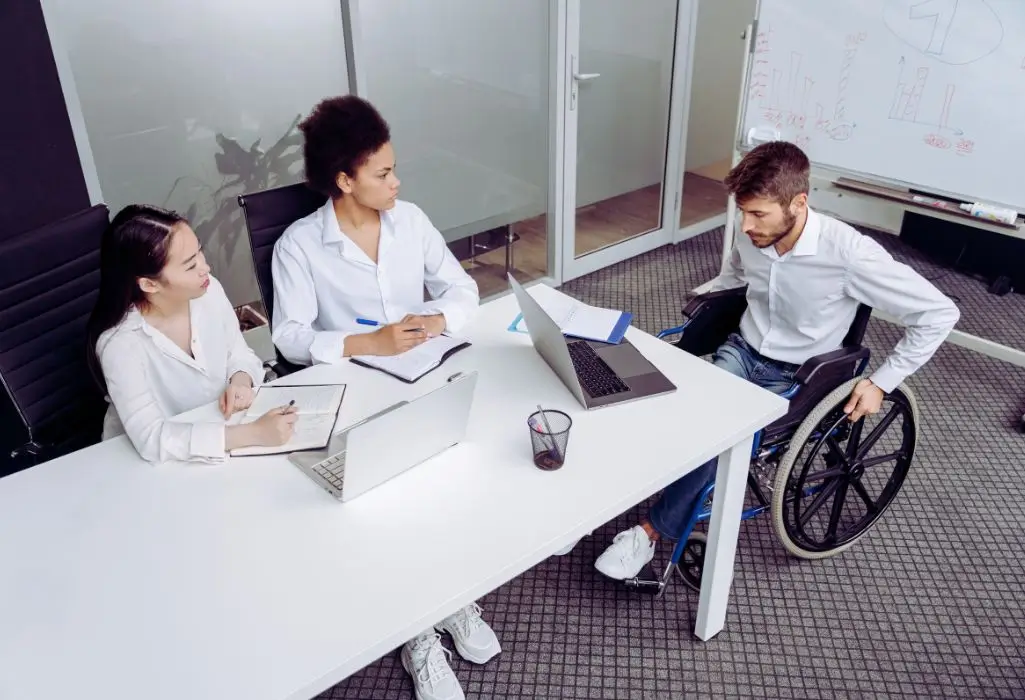 Na zdjęciu widać spotkanie rekrutacyjne – przy białym stole siedzą dwie kobiety prowadzące rozmowę oraz mężczyzna na wózku inwalidzkim, który uczestniczy w procesie rekrutacyjnym. Wszyscy mają przed sobą laptopy i notatniki, co podkreśla profesjonalny charakter spotkania. Atmosfera zdjęcia łączy w sobie skupienie, otwartość i równość – pokazuje, że pytanie jak zatrudnić osobę niepełnosprawną nie dotyczy tylko formalności, ale także tworzenia przestrzeni, w której każdy kandydat jest traktowany z szacunkiem i ma równe szanse.