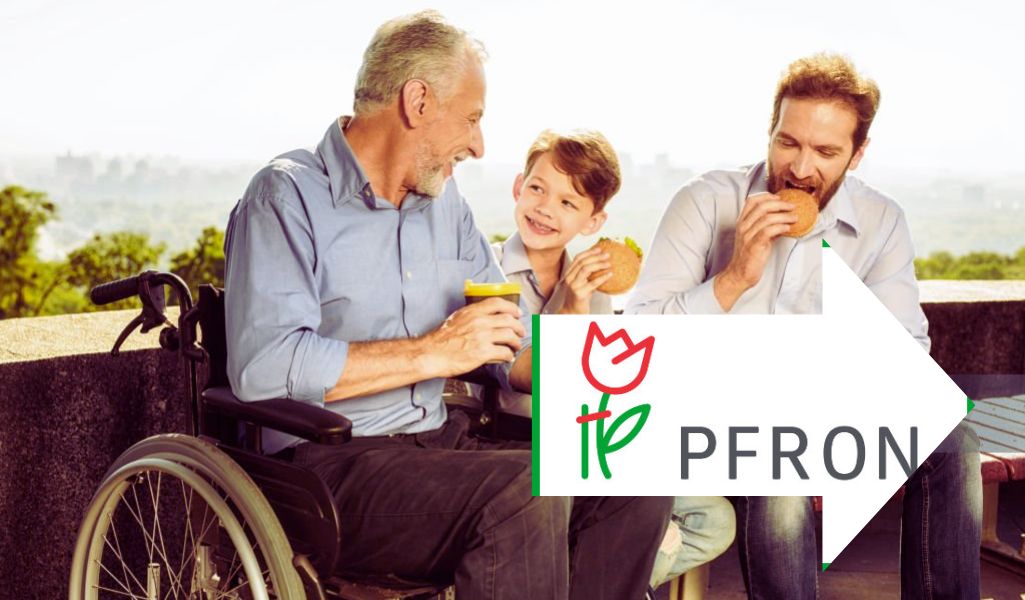 Ojciec na wózku inwalidzkim spędza czas z dzieckiem i drugim dorosłym na świeżym powietrzu. Wspólne jedzenie i uśmiech symbolizują wsparcie oraz dofinansowanie do opieki nad dzieckiem z PFRON.